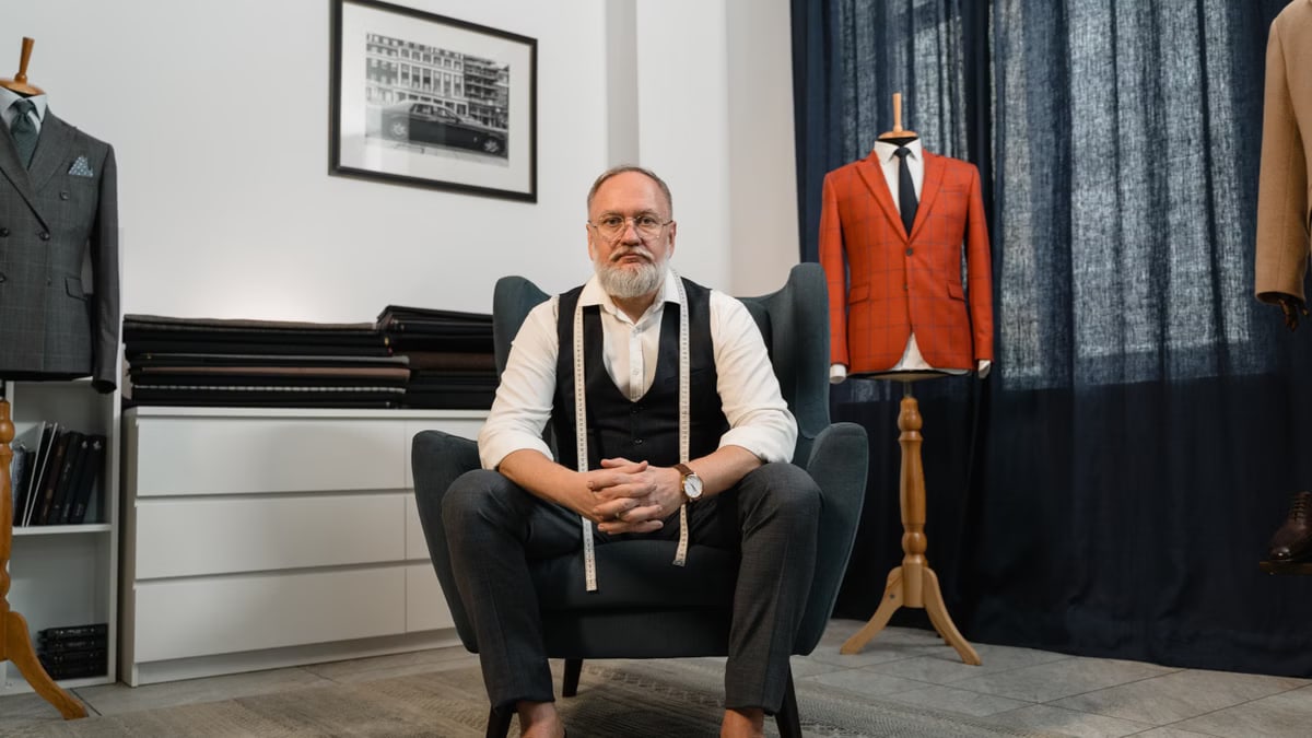 Elderly tailor sitting in an elegant shop with suits and mannequins.